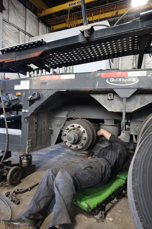 Mechanic on a creeper under a car-hauler trailer working the hub, air bag, and suspension.